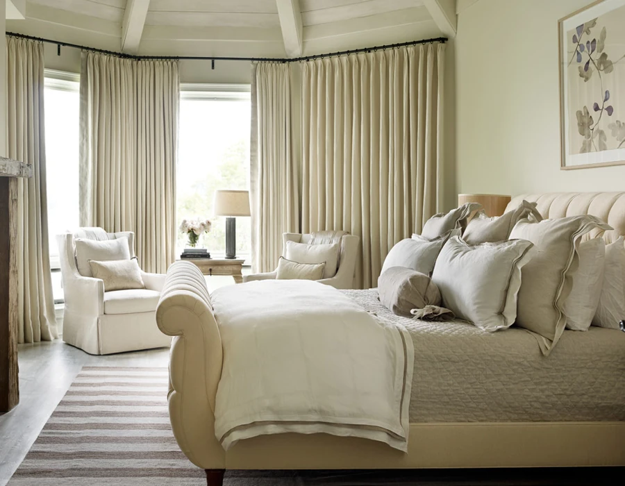 cream and ivory bedroom with pleated curtains, a bed with a tufted headboard, and armchairs by the windows