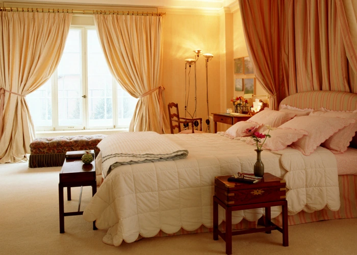 Softly draped peach curtains hang from a canopy bed and large window in a luxurious bedroom.